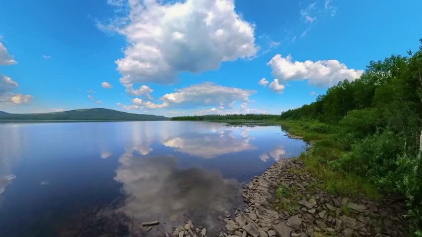 panoramic view of a calm lake surrounded by green trees and distant hills under a bright blue sky with fluffy white clouds, showcasing a perfect reflection on the water