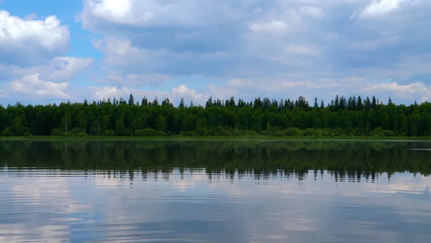 A serene view of a calm lake in the Southern Urals region of Russia on a sunny day. The clear water reflects the lush green forest on the distant shore and the blue sky with white, fluffy clouds above