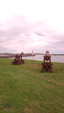 Two antique cannons are positioned on a grassy hill overlooking a body of water. In the distance, a lighthouse stands near a pier. The sky is overcast with soft clouds, and the overall scene suggests a historical coastal defense site. Shot on: November 18, 2025 at 12:08 PM. Location: Saint-Pierre, Saint-Pierre-et-Miquelon, France.