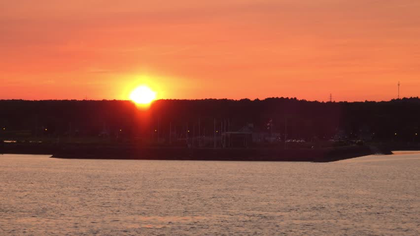 The sun is setting over a body of water, casting a warm orange glow across the sky. In the foreground, the water is calm. A dark silhouette of land is visible in the mid-ground, featuring a harbor with numerous sailboats and some buildings. The horizon is lined with trees, and a few distant structures and power lines are discernible. Shot on: November 18, 2025 at 12:08 PM. Location: Portneuf, Québec, Canada.