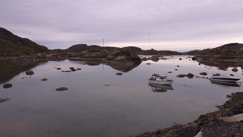 A serene scene of several boats, including a white cabin cruiser and a smaller vessel, gently bobbing on the still water of a bay. The surrounding landscape is characterized by rocky, undulating hills and a few scattered buildings, one with a red roof. The sky is overcast, casting a soft, diffused light over the tranquil setting. The water reflects the sky and the surrounding terrain, creating a peaceful atmosphere. Shot on: November 18, 2025 at 12:09 PM. Location: Sisimiut, Kalaallit Nunaat. Greenland
