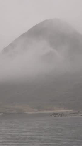 A serene view of a misty coastline. Fog partially obscures a rugged cliff face that slopes down towards the water. The water