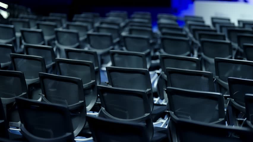 Empty rows of modern black mesh conference chairs fill a large auditorium, chrome frames reflecting blue stage lighting, awaiting attendees before a business presentation or keynote
