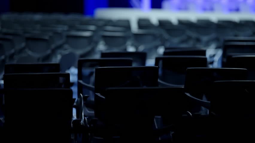 Rows of empty conference chairs in a dimly lit auditorium with cool blue stage lighting, capturing the quiet anticipation before a presentation begins in a modern event venue