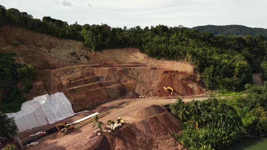 Professional aerial drone footage documenting active extraction operations in Busuanga area, Coron Island, Palawan, Philippines. Smooth forward flight reveals industrial-scale open-pit site with heavy machinery removing minerals from terraced mountain excavation surrounded by tropical rainforest.
Multiple yellow excavators and earthmoving vehicles operate across different levels, with visible access roads and stockpiled materials. White protective shelter covers storage area in foreground. Terraced benches cut into hillside show systematic extraction process with red-orange soil layers exposed through industrial activity. Palm trees dot the operational zone, providing scale reference against massive earthworks.
The site features clearly defined terracing with stepped walls rising toward jungle-covered peaks. Mountains blanketed in dense vegetation form dramatic background under overcast sky. Processing area at ground level shows organized layout with vehicles, material piles, and infrastructure supporting continuous operations.
This footage captures real working conditions at Southeast Asian mineral extraction facility, showing scale of industrial operation, terrain modification, and coexistence of heavy industry with natural environment. Perfect for documentary projects about extractive industries, natural resource management, Philippine economic development, remote industrial operations, or environmental impact studies.
High-resolution cinematography with stable gimbal movement provides broadcast-quality content suitable for news coverage, corporate presentations, educational materials, or editorial pieces discussing resource extraction, infrastructure development, geological surveys, occupational safety, or sustainable management practices in developing regions.