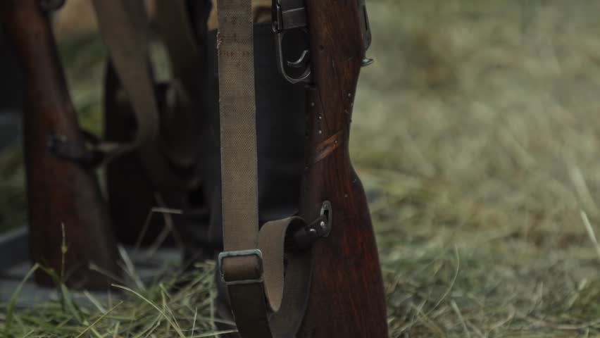 Red Army private soldier holds Mosin rifle, standing guard near display of weapons and army tents. Reenactment portrays field life of Great Patriotic War.