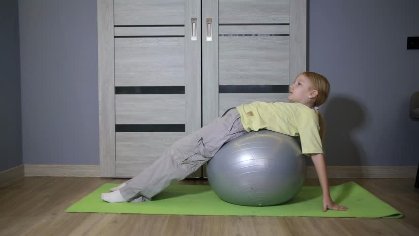 Young caucasian girl with light hair and yellow t-shirt performing leg raise exercise on grey fitness ball. Child lies on stability ball on green yoga mat during indoor workout at home.