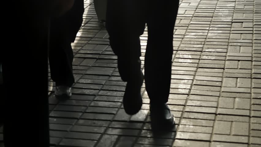 Low-angle glimpse of pedestrians walking across a sunlit brick sidewalk, their legs casting long shadows while glass surfaces mirror their movement in a quiet urban scene