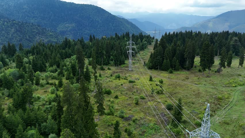 High voltage power line cutting through a thick pine forest in a mountain valley, overhead view of transmission towers and cables against a cloudy sky, where energy meets the rugged wilderness