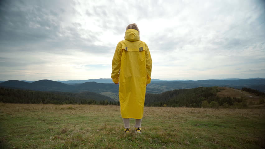 Person in yellow raincoat on hill overlooking vast moorland under moody sky arms raised in quiet celebration, facing wind, soft grass underfoot, rolling ridges
