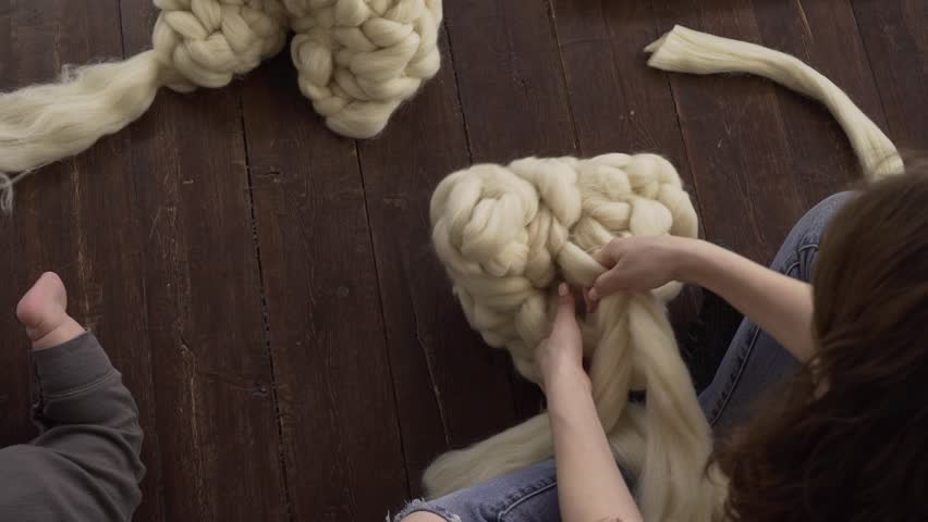 A young woman sitting on the floor in her apartment, weaves a product made of thick merino wool by her hands and her baby crawls next to her. Female freelance on maternity leave.