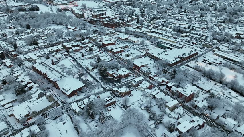 Aerial top down shot of dawn scene in snow- and ice-covered American town, with rooftops, streets and trees coated in fresh winter snowfall. Blue cold morning in neighborhood of U.S. Rising shot.
