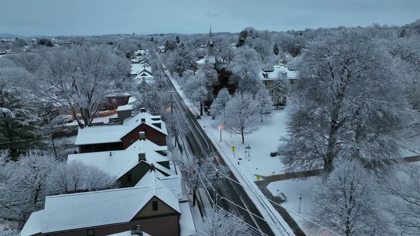 Aerial flyover american town named Lititz, Pennsylvania in winter dawn, showing snow-covered main road, historic church steeple and quiet houses surrounded by frosted trees. Wide shot. Snowy roofs.
