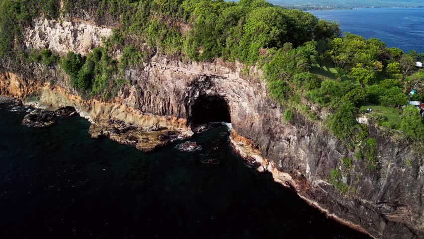 Aerial drone footage showing Bali limestone cliffs rising above deep blue sea with a natural sea cave, lush tropical trees on top and calm ocean movement shaping the rugged island shoreline