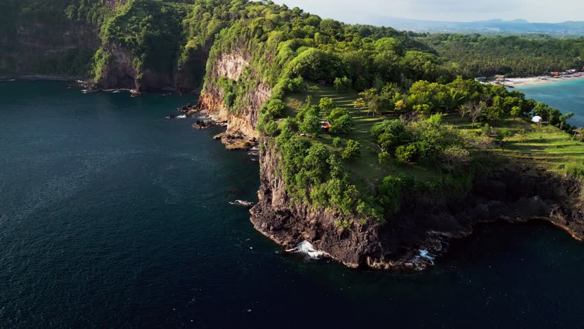 Aerial drone footage of a Bali tropical coastline showing volcanic rock cliffs, forested hills, coastal headlands, and clear ocean water, with a secluded beach and calm coastal surroundings.