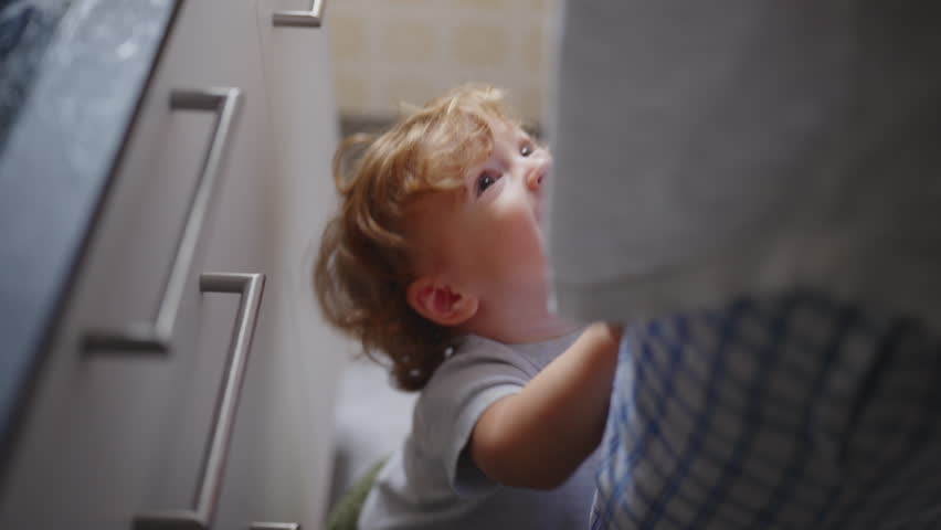 Toddler crying for attention and pushing mother away from kitchen counter before she finally lifts him into her arms, candid emotional family moment at home