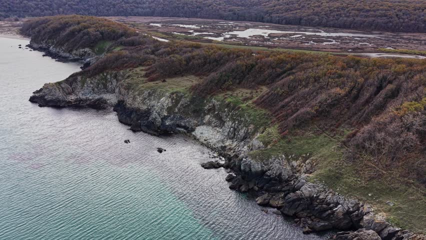 Stunning aerial perspective showcases a rugged coastline meeting tranquil waters, surrounded by colorful autumn foliage and a winding path through the landscape.