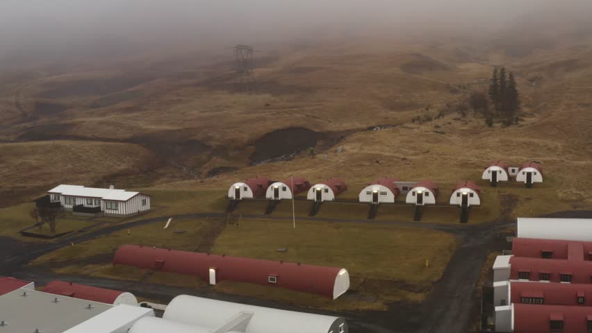 Aerial drone view of red and white Quonset huts aligned on green lawns in Hvalfjörður Iceland with foggy hills and rural landscape in the background during overcast weather