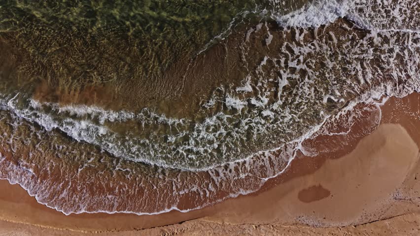 Waves roll onto a warm sandy beach, creating a beautiful contrast with the clear blue water under bright sunlight. The shoreline curves invitingly.