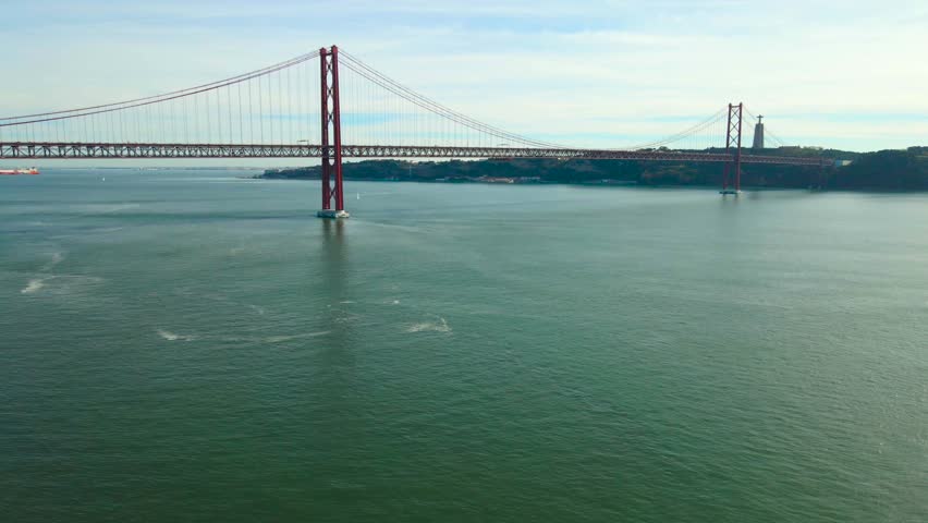 Drone shot over the Tagus River showing the red Ponte 25 de Abril suspension bridge with traffic and the Sanctuary of Christ the King monument in sunny Lisbon.