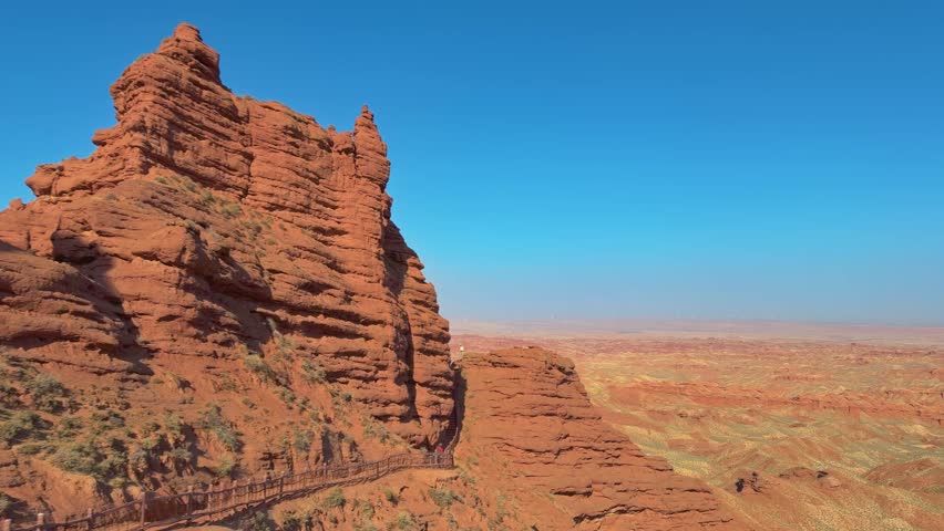 Pingshan Lake Grand Canyon, Zhangye, Gansu Province, China - Towering Red Rock Cliffs Rise Sharply Beside a Narrow Walkway, Overlooking a Vast, Eroded Canyon Under a Clear Blue Sky - Pan Right Shot