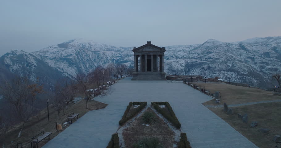 Aerial drone footage moving forward and upward around Garni Temple in Armenia during blue hour after sunset. The temple gradually disappears from the frame as the camera ascends, revealing snow-covered mountains and surrounding landscape under evening blue light. Filmed around 21:00 in winter conditions. Calm atmospheric scene suitable for travel, tourism, documentary, and commercial projects.