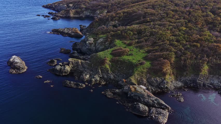 This aerial view showcases a serene coastal landscape featuring a rocky shoreline, lush green patches, and deep blue water. Nature