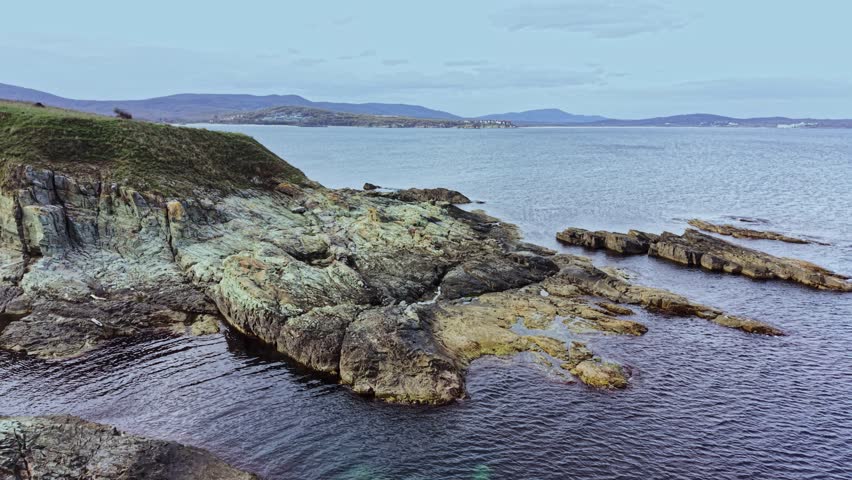 This aerial perspective showcases a rugged coastline with rocky formations, calm waters, and distant hills under a clear sky, capturing the serene beauty of the landscape.