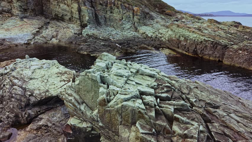 An aerial view shows the rugged rocks and calm waters along the coastline. The landscape features various rock formations and gentle waves reflecting the sunlight.
