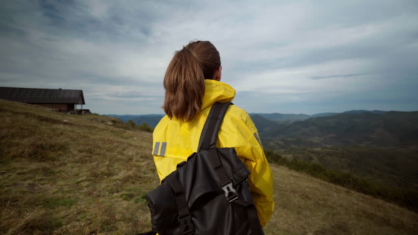 Woman in yellow raincoat looking across valley from ridge, trail scout surveys weather and terrain while shading eyes, rustic cabin and moody clouds frame