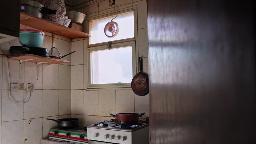 Rustic home kitchen interior with gas stove simmering a pot, hanging pan, tiled walls and shelves, soft daylight through window. Authentic everyday cooking background.