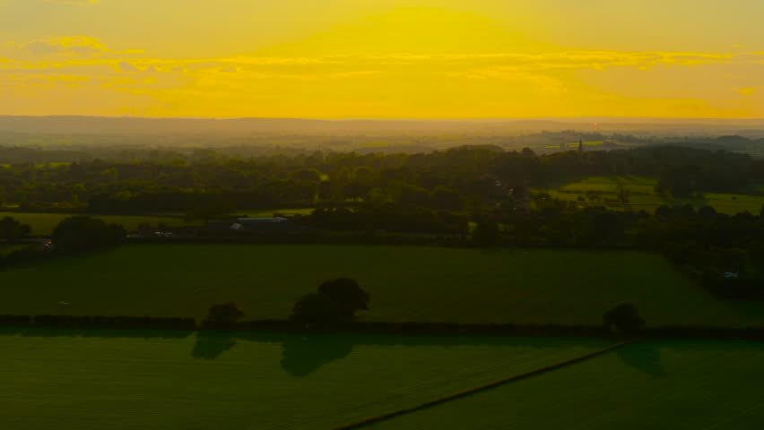 Vast farmland stretching under golden sunset over distant hills. Evening sunlight painting fields and forests in warm tones. Agricultural valley glowing under radiant yellow sky