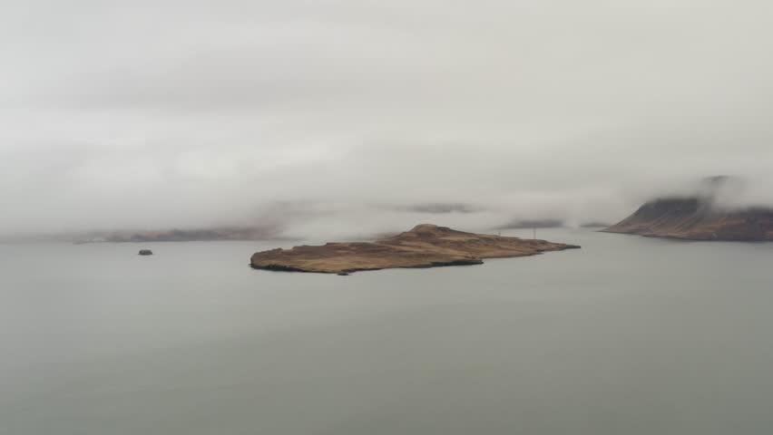 Clouds rolling over isolated island in calm misty bay of Hvalfjörður Iceland surrounded by still water and dramatic low fog under an overcast sky