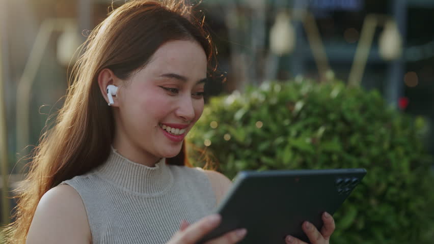 Portrait of happy Asian woman with earphones watches video via digital tablet in green park. Positive Korean lady looks at mobile device screen in downtown district on windy day