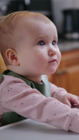 A cheerful infant girl sits in a high chair, eats from a spoon held by her mother, and claps her hands happily. Vertical video.