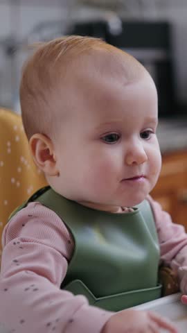 A cheerful infant girl sits in a high chair and smiles while being fed with a spoon. Vertical video.