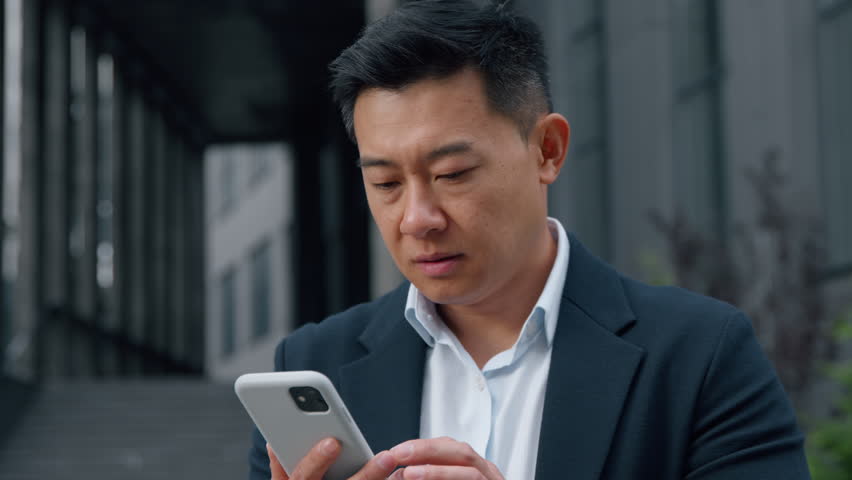 Middle-aged Asian businessman in city with phone sad Korean business man with mobile app failure upset worried looking in smartphone Chinese Japanese male entrepreneur outdoors using gadget problem