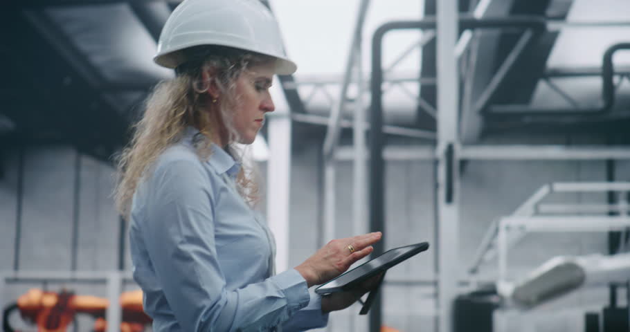 Professional Woman Engineer in Safety Helmet Checking Information on Tablet While Walks in Fully Automated Factory With Robotic Machinery. Concept of Modern Industry and Digital Production Control.