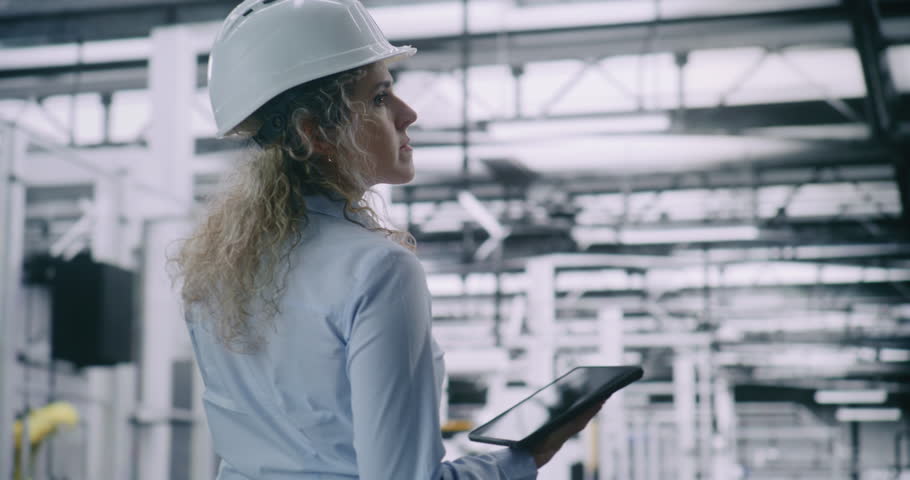 Female Technical Supervisor in Safety Gear, Overseeing Complex Machinery on Smart Factory. Woman Utilizes Mobile Device to Ensure Operational Compliance and Technical Standards Adherence. Back View.