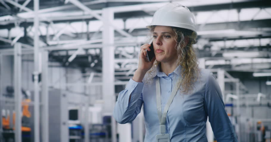 Blonde Curly haired Engineer in White Safety Helmet Walks Through Factory Floor While Talking on Smartphone, Coordinating Operations in Automated Facility. Concept of Communication and Management.