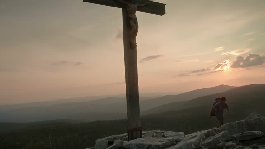A person admires the sunset while standing by a wooden cross. The view captures mountains and clouds during golden hour. Nature surrounds the area.