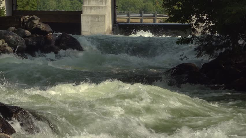 A close-up of white water flow rapids flowing under a sturdy stone bridge with a dam. Rushing water and large boulders along the bank make for great dynamic landscape.