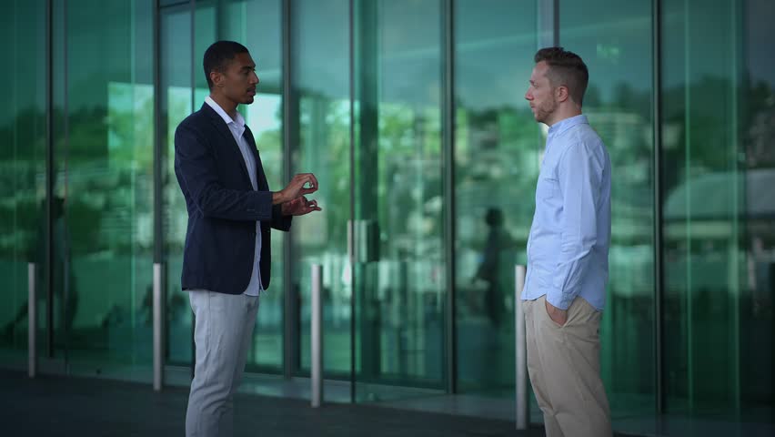 Two young diverse male business people chatting together standing in front of modern corporate glass building