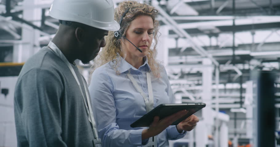Two Specialists Solve Technical Issue in Vast Industrial Plant. Woman Points Area Concern on Factory Floor, Using Tablet For Data Reference. Problem Solving and Operational Excellence in Industry 4.0.