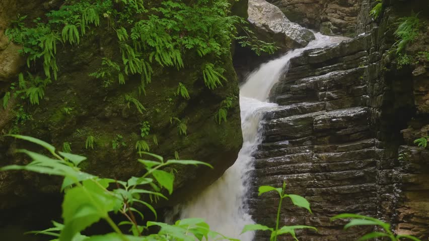footage of a beautiful waterfall flowing through a natural rock gorge with vibrant green foliage. The water cascades over unique, naturally formed stone steps.