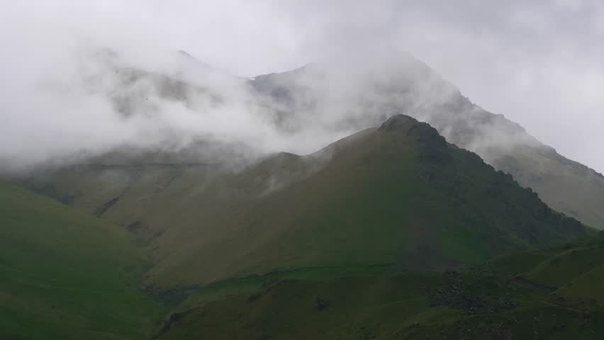 A slow drone movement captures the serene beauty of the Kara-Syrt Ridge in Dagestan, part of the greater Caucasus Mountains range. Lush green slopes and peaks are shrouded in swirling mist and clouds,