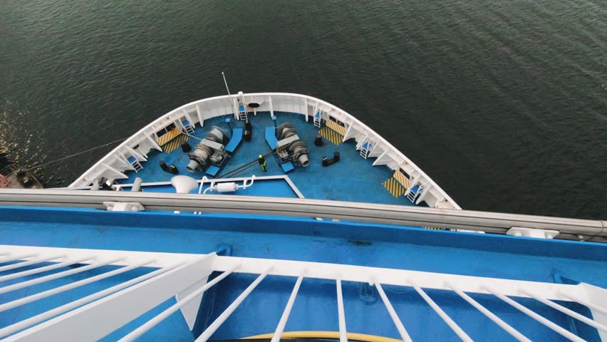 View from above of a ferry bow with blue deck and mooring equipment cutting through calm water, maritime transport scene, modern passenger ship, no people, overcast daylight