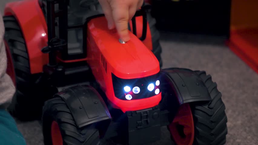 Close-up footage of a child’s hand engaging with a red toy tractor. The shot focuses on the tactile play, showing the child’s interaction with the agricultural vehicle model. The toy features large treaded tires and realistic farming machinery details.