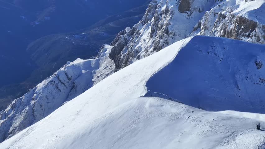 A stunning high-altitude view of a rugged mountain range covered in a fresh layer of snow. The late afternoon sun casts long, dramatic shadows across the slopes and ridges, highlighting the sharp textures of the terrain against a deep blue sky.