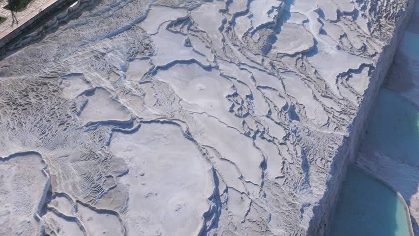 Aerial drone footage of the white travertine terraces of Pamukkale in Turkey, filled with natural thermal water pools. The bright mineral formations create a striking contrast with the surrounding landscape under soft sunlight. This unique geological site, formed by calcium-rich hot springs, is one of Turkey’s most iconic natural landmarks and a UNESCO World Heritage location, ideal for travel, tourism, documentary, and nature projects.